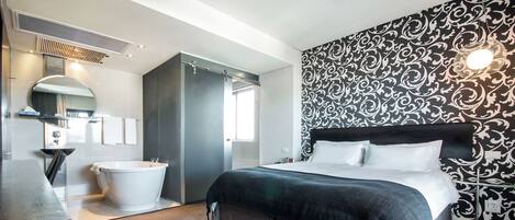 A modern hotel room with a large bed, a bathtub, a vanity with a mirror, and a patterned wall.