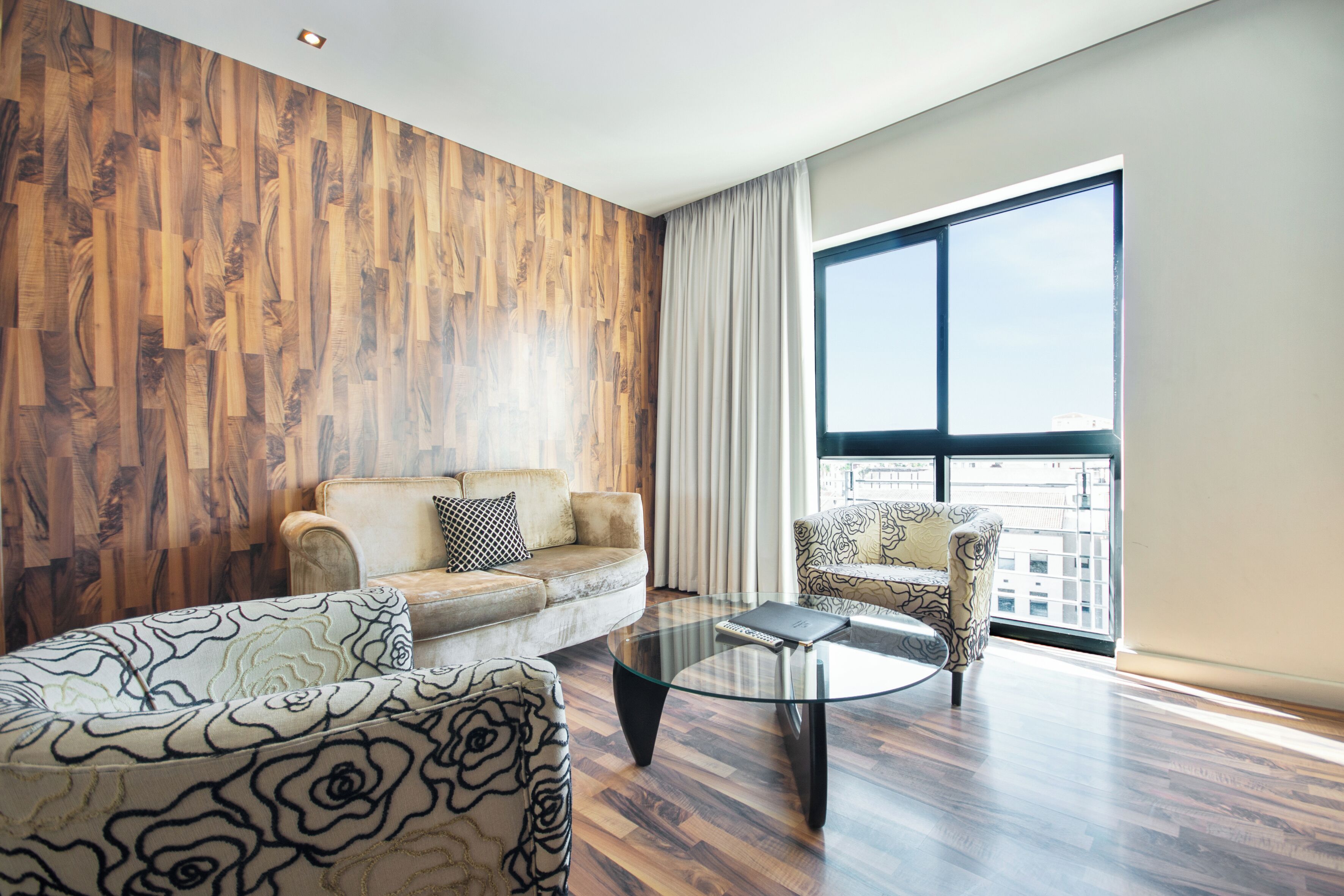 A modern living room with a wooden wall, a sofa, a patterned armchair, a glass coffee table, and a large window with a view.