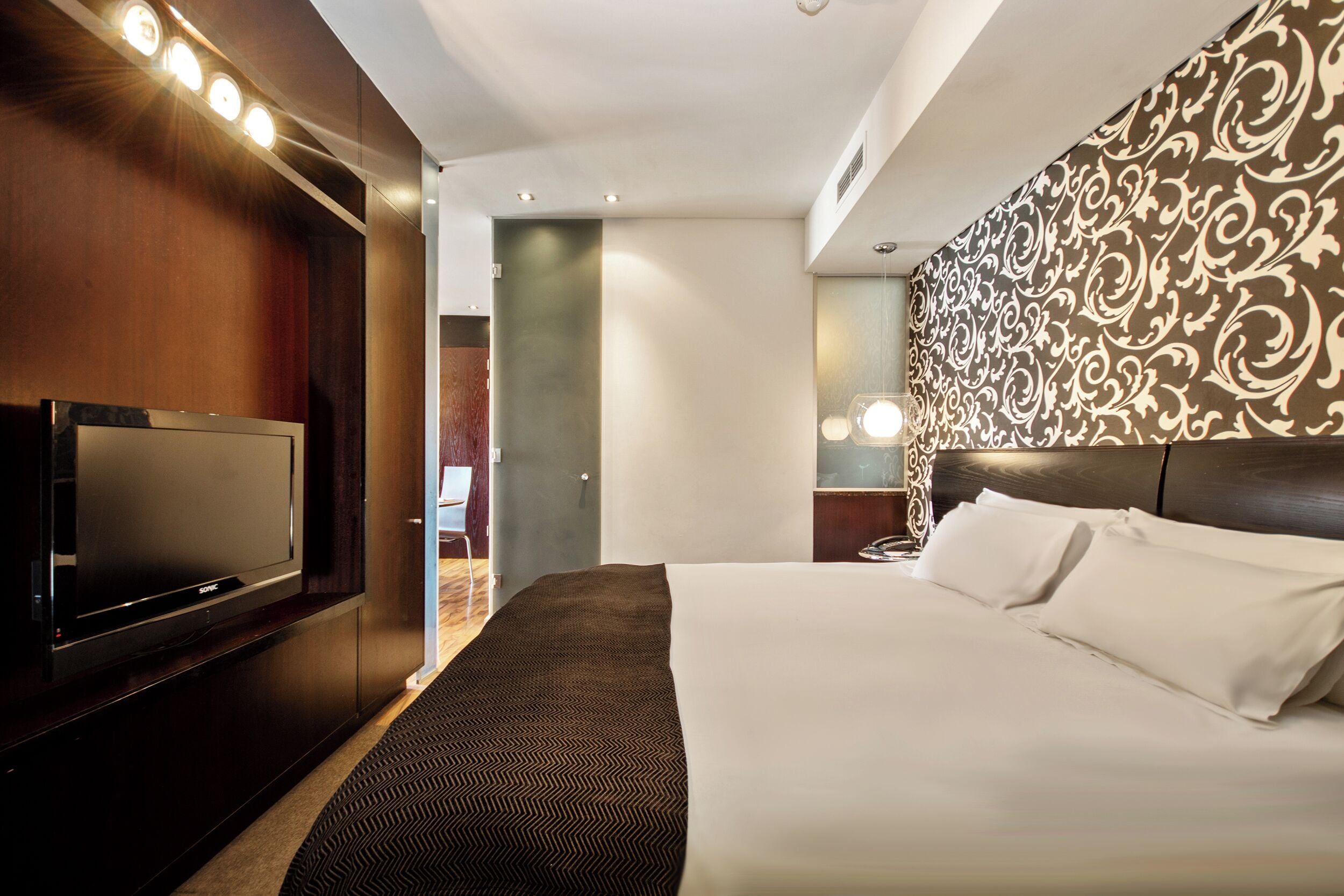 A hotel room with a bed, a TV, a patterned wall, and a nightstand with a lamp.
