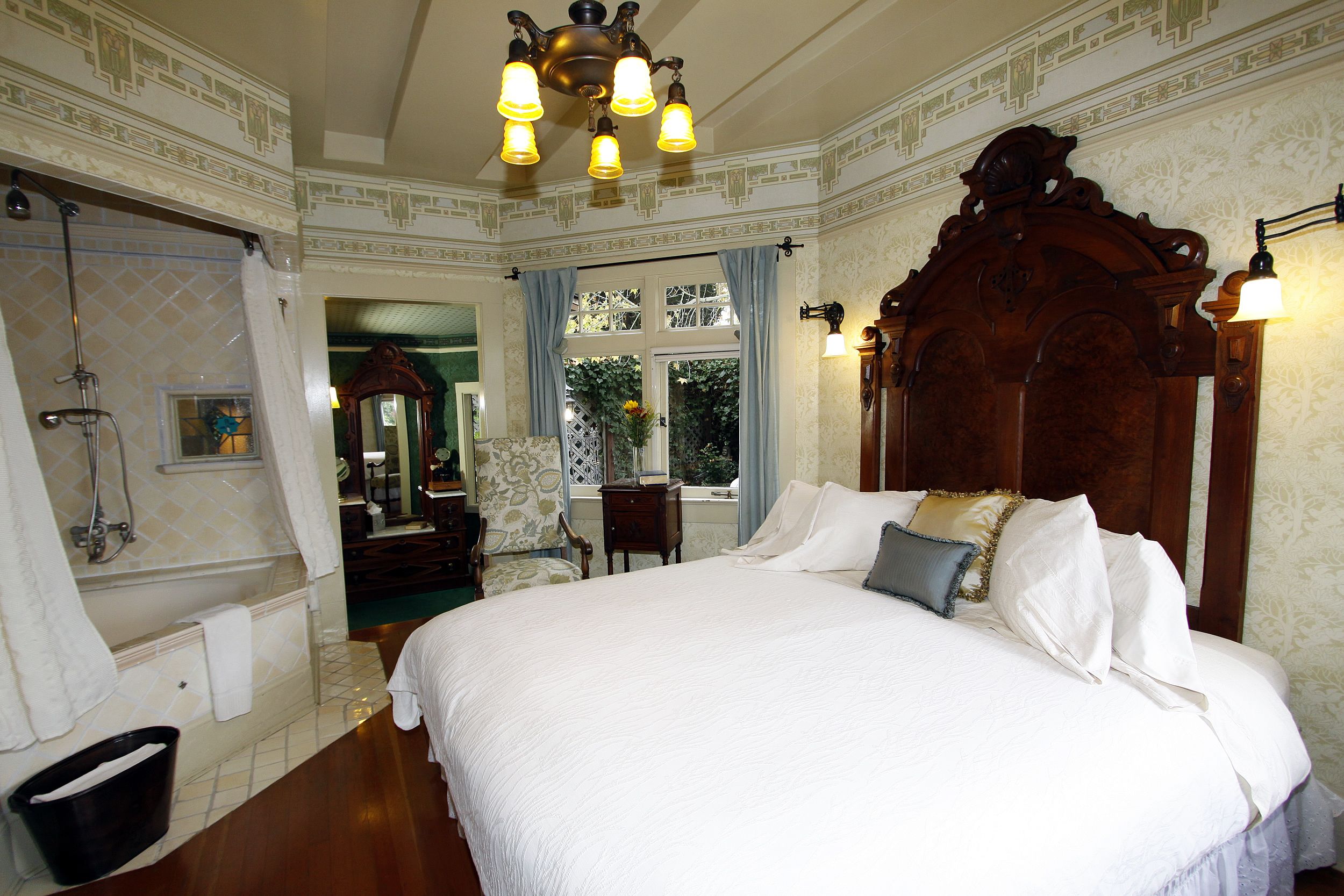 A large bed with a wooden headboard, a bathroom with a shower, and a window with curtains.
