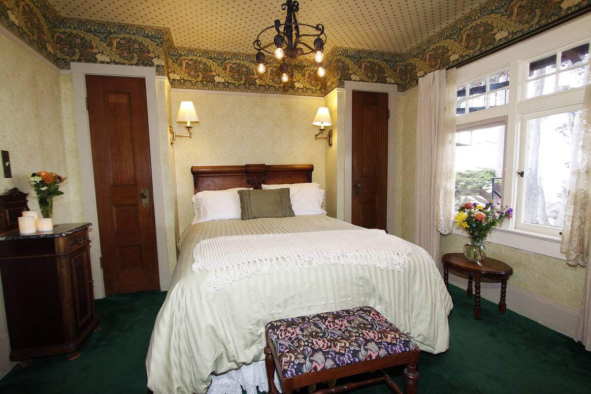 A bedroom with a large bed, two wall-mounted lamps, a wooden bench, a nightstand, and a window with curtains.