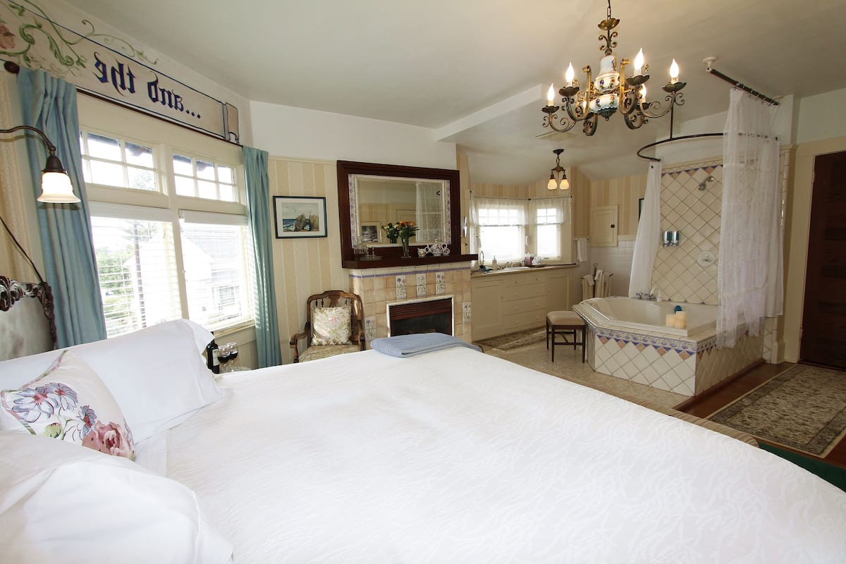 A bedroom with a large bed, a sitting area with a chair, a fireplace, and a bathroom with a bathtub and shower.