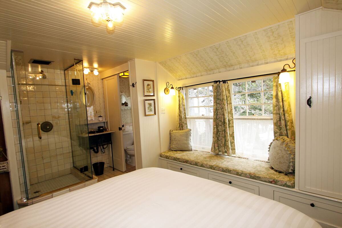 A bedroom with a large bed, a window seat with curtains, a shower area, and a bathroom with a toilet and sink.