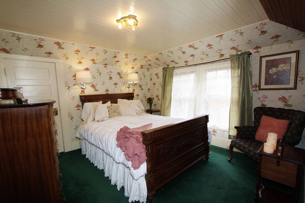 A bedroom with a wooden bed, floral wallpaper, a green carpet, a chair, and a bedside table with a lamp.