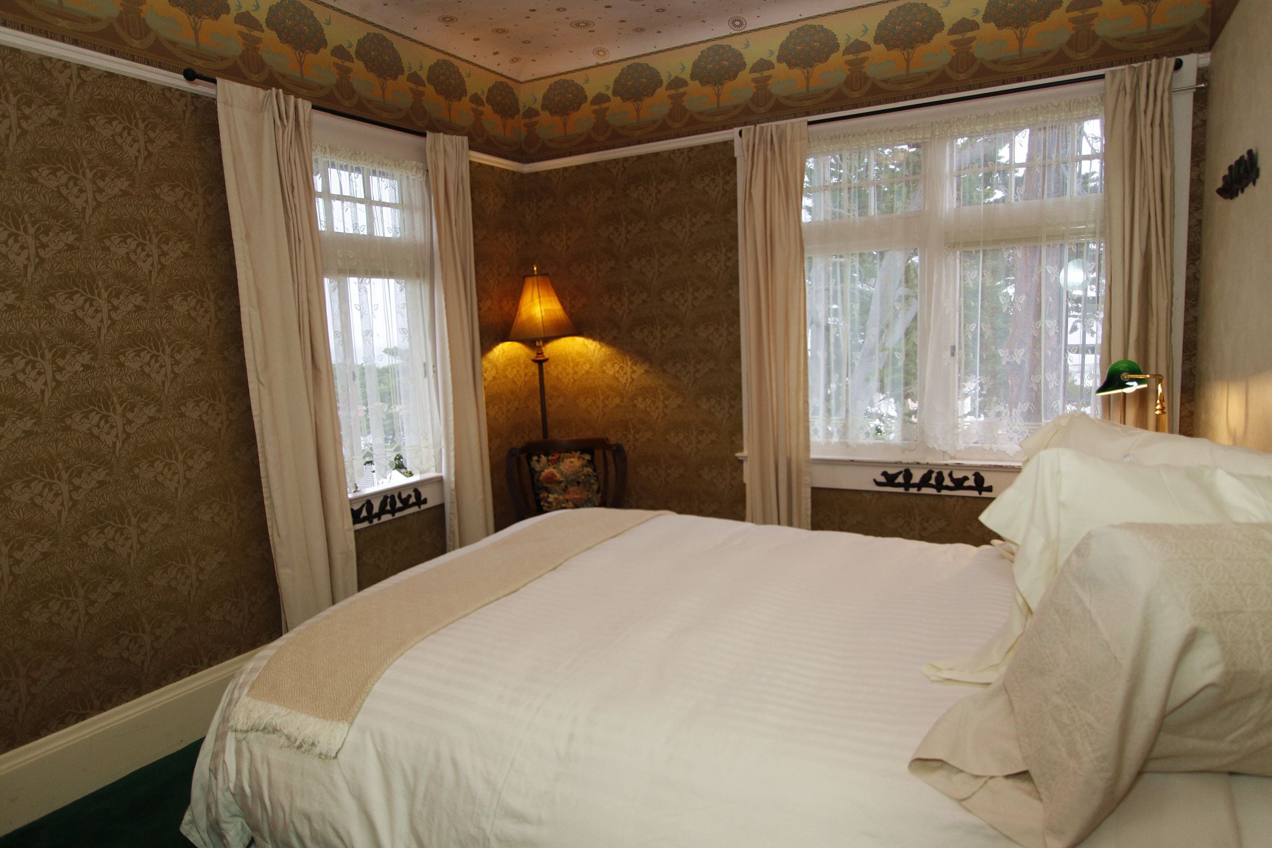 A bedroom with a large bed, two windows with curtains, a lamp, and a chair.