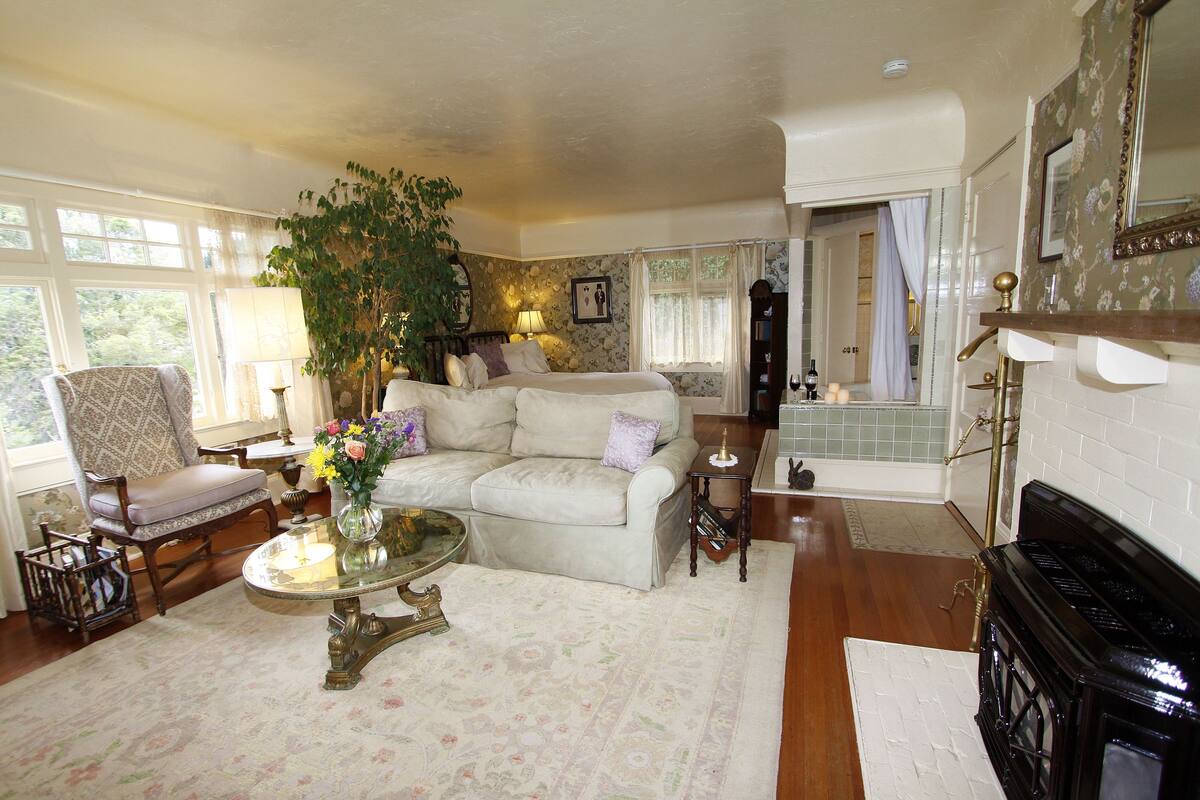 A cozy living room with a sofa, armchair, and a coffee table. There is a fireplace, a large window, and a plant.