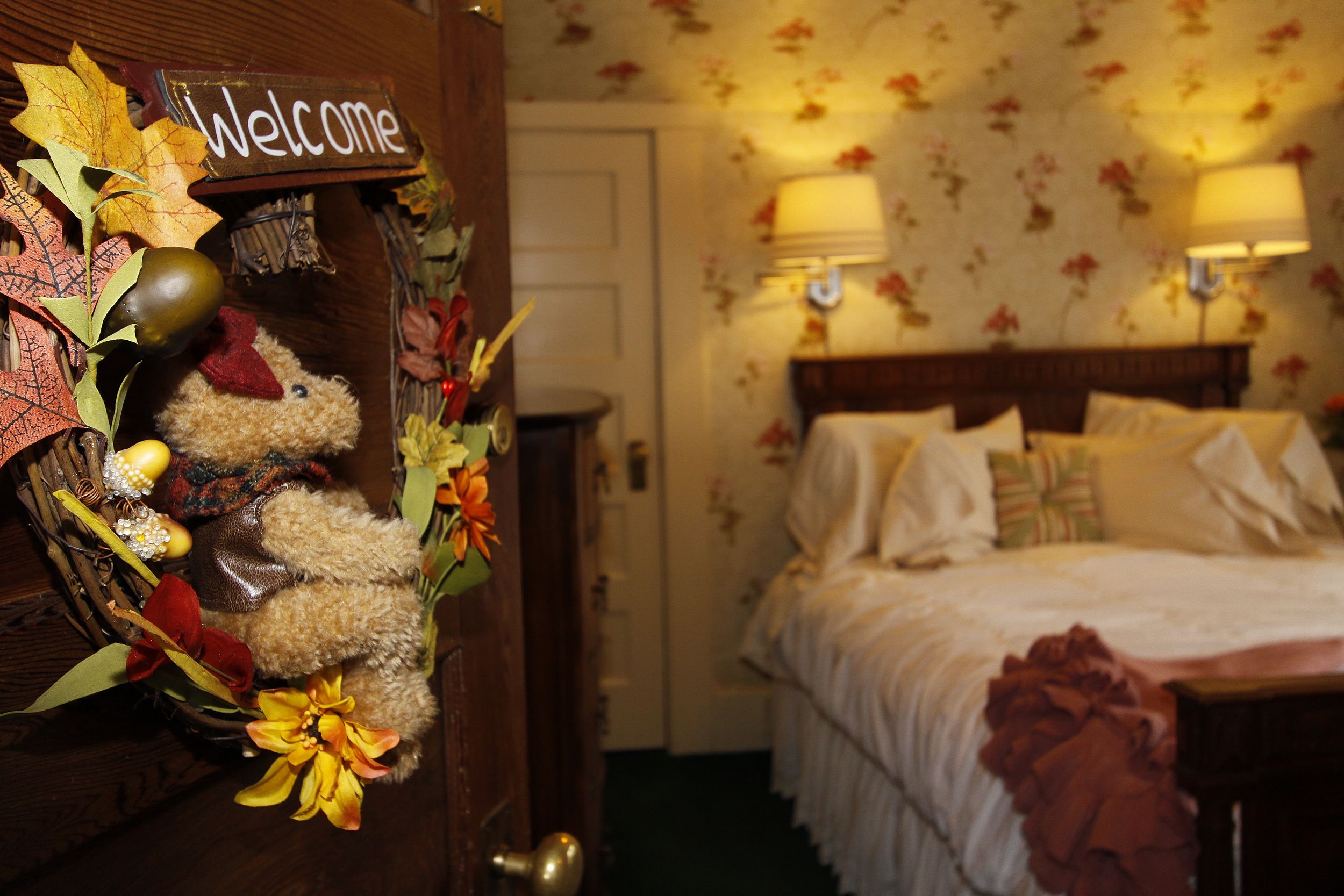 A hotel room with a bed, two bedside lamps, a floral wallpaper, and a welcome sign with autumn leaves and a teddy bear.