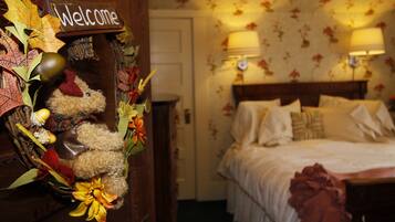 A hotel room with a bed, two bedside lamps, a floral wallpaper, and a welcome sign with autumn leaves and a teddy bear.