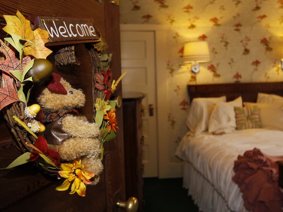 A hotel room with a bed, two bedside lamps, a floral wallpaper, and a welcome sign with autumn leaves and a teddy bear.
