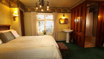 A hotel room with a large bed, a bedside table with a lamp, a window with curtains, a bathroom with a sink and mirror, and a door leading to another room.