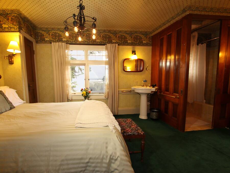 A hotel room with a large bed, a bedside table with a lamp, a window with curtains, a bathroom with a sink and mirror, and a door leading to another room.