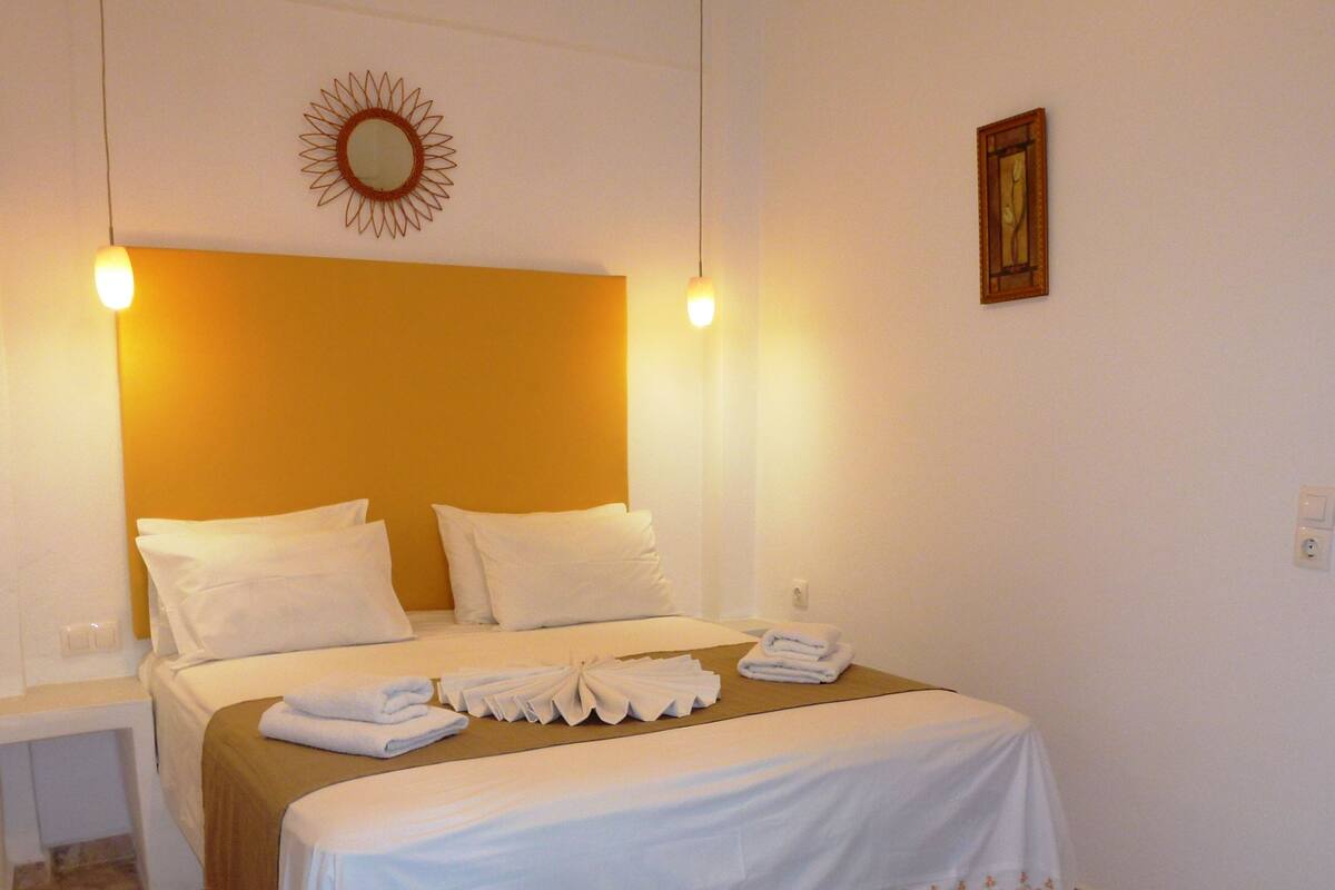 A neatly made bed with white linens and a decorative border, a bedside table with a lamp, and a wall-mounted mirror.