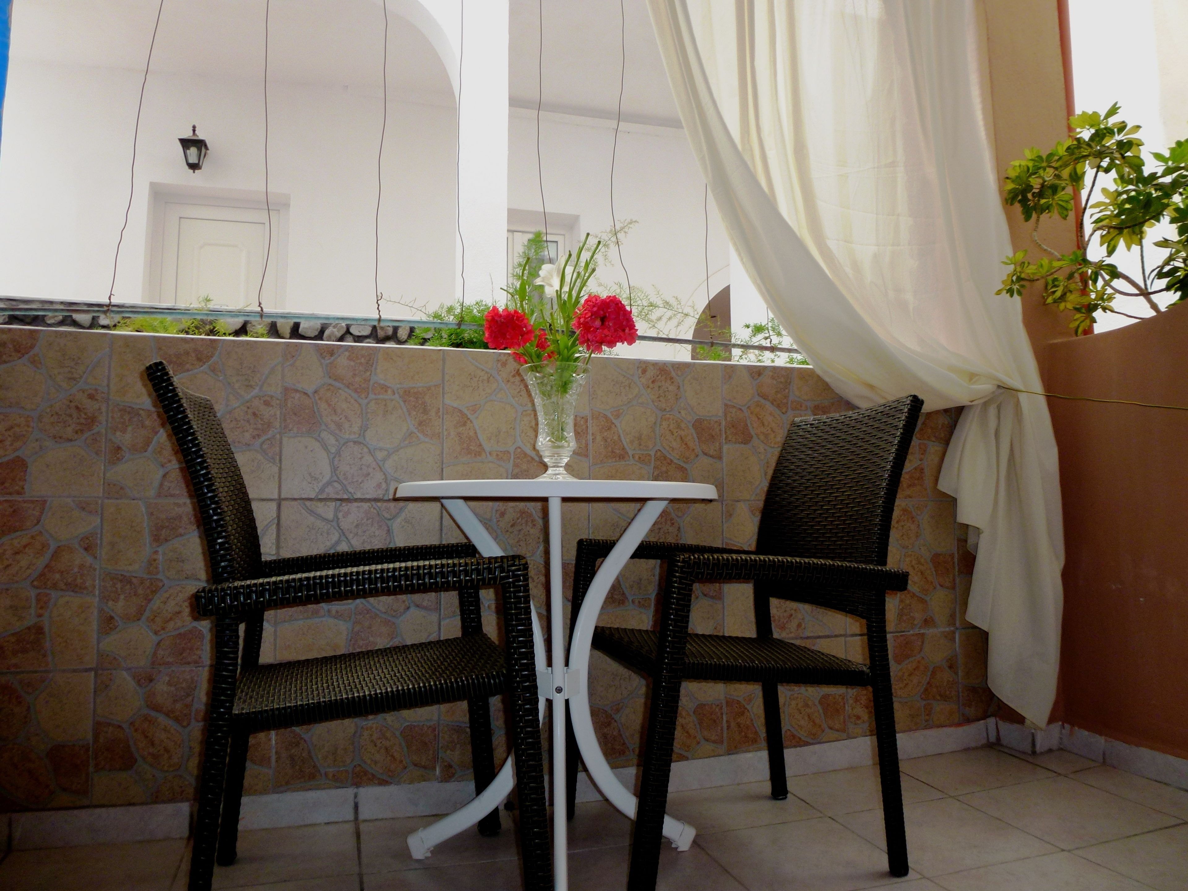 A small outdoor seating area with two wicker chairs and a white table, a vase with red flowers, and a stone wall backdrop.