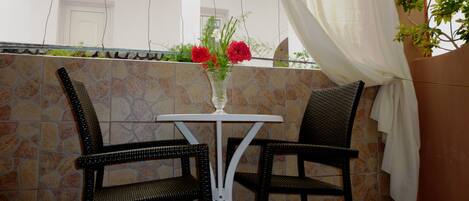 A small outdoor seating area with two wicker chairs and a white table, a vase with red flowers, and a stone wall backdrop.