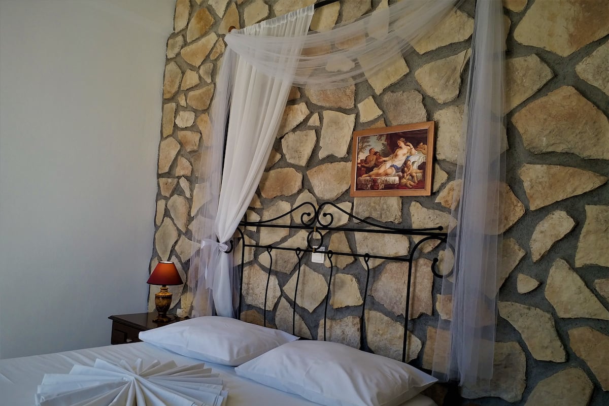 A neatly made bed with white linens and pillows, a stone wall background, a framed painting, and a bedside lamp.