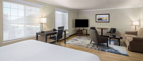 Una habitación de hotel con una cama grande, un escritorio con silla, una mesita con una silla, un sofá, un televisor y una ventana con vistas al exterior.