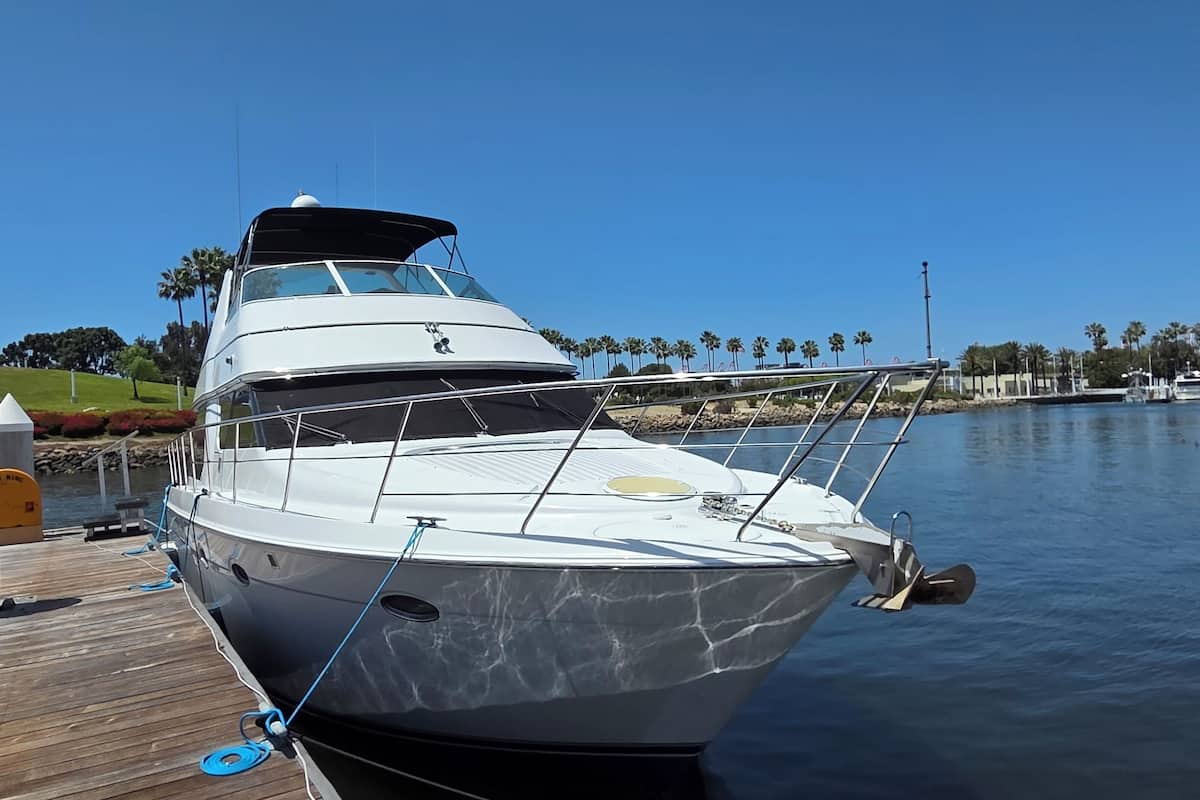 A yacht docked at a marina with a wooden pier, a lighthouse in the background, and palm trees along the shore.