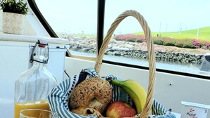 Una mesa con un cesto de mimbre que contiene bagels, frutas y una botella de jugo, con vistas a un paisaje a través de una ventana.