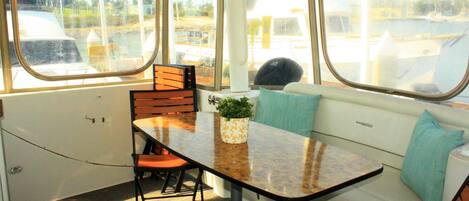 A sunlit interior with a table, chairs, and a sofa by a large window overlooking a marina.