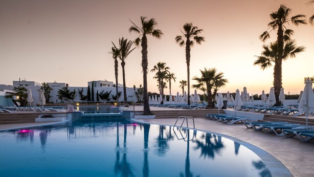 A pool area with lounge chairs and umbrellas, surrounded by palm trees and buildings. - Aparthotel Marinda Garden (Ciutadella de Menorca)