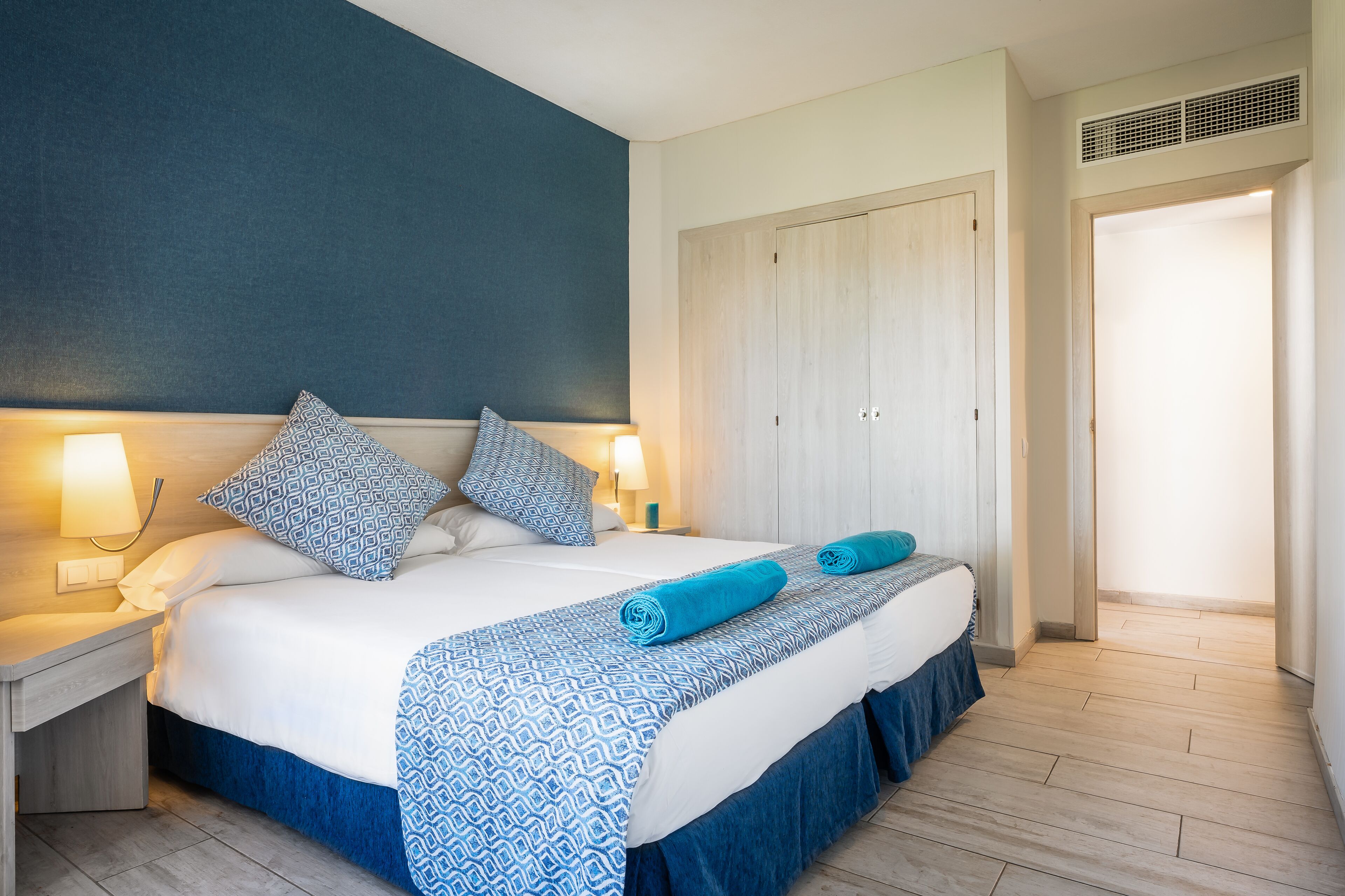 Una cama bien hecha con una colcha estampada, dos almohadas y una toalla enrollada. Se observa un mesita de noche con una lámpara y una luz empotrada en la pared. En el fondo, un armario con puerta es visible.