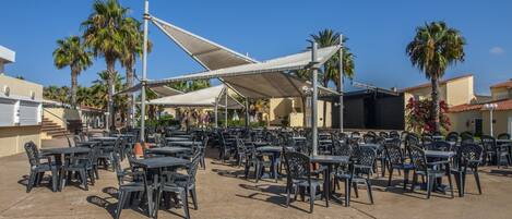 Een terras met tafels en stoelen, omgeven door palmbomen en gebouwen.