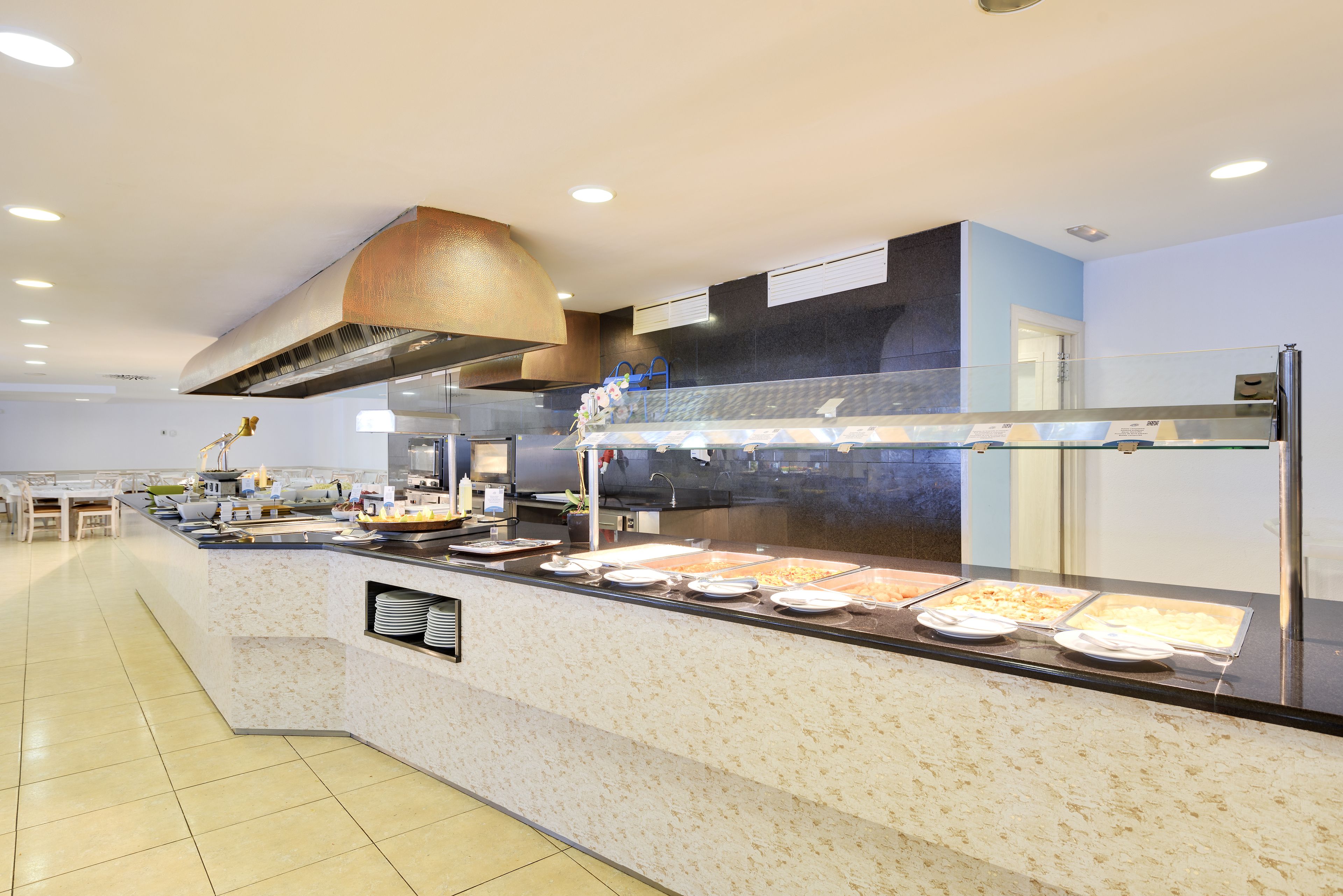 Een buffetgedeelte met verschillende food stations, een grote ventilatiekap erboven en een zithoek met tafels en stoelen op de achtergrond.