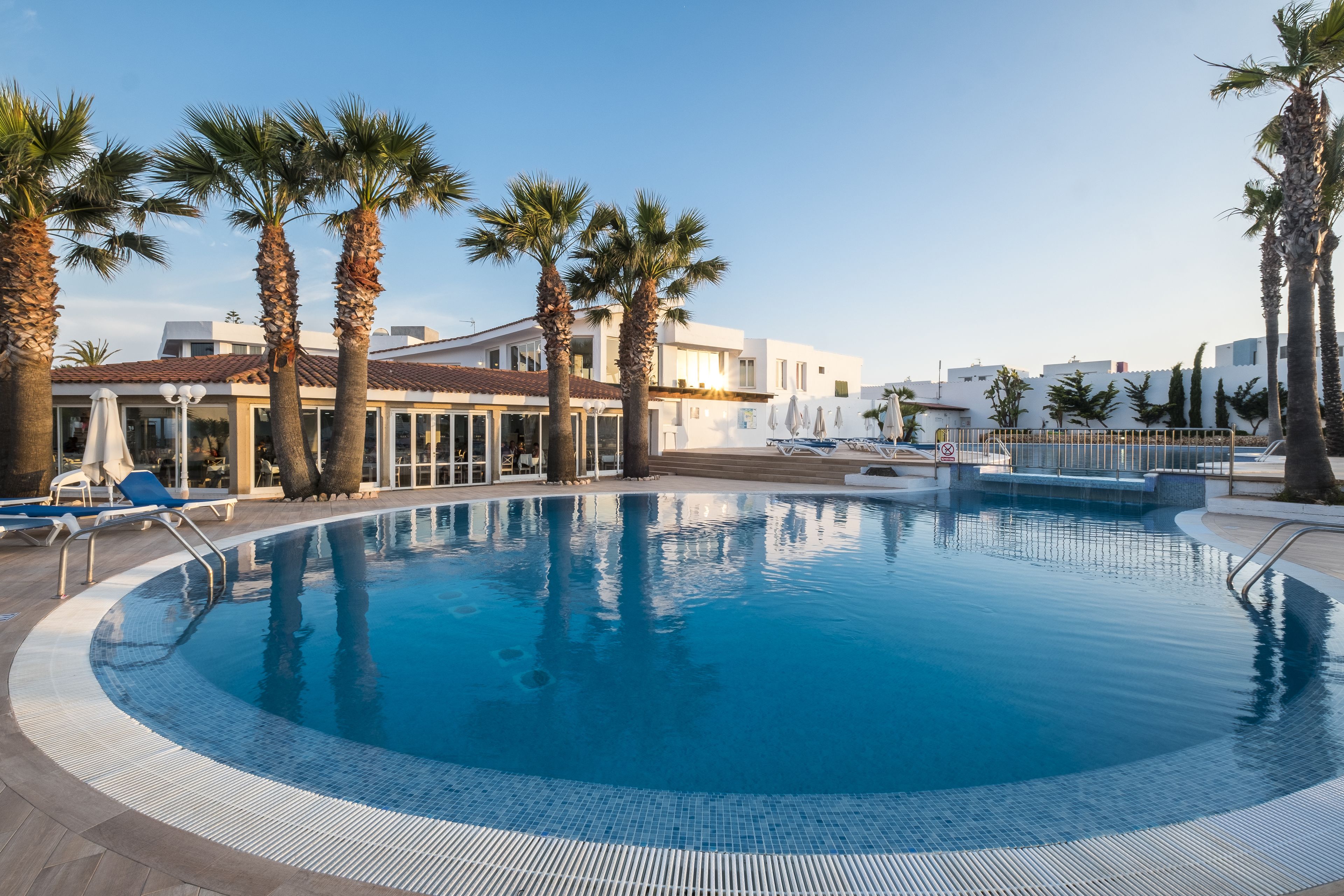 Piscina con palmeras y sillas de descanso, rodeada por un edificio con grandes ventanales y una terraza con baldosas.
