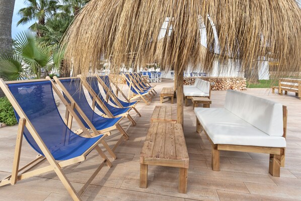 A row of blue lounge chairs and white sofas under a thatched umbrella, with wooden benches and a stone wall in the background.