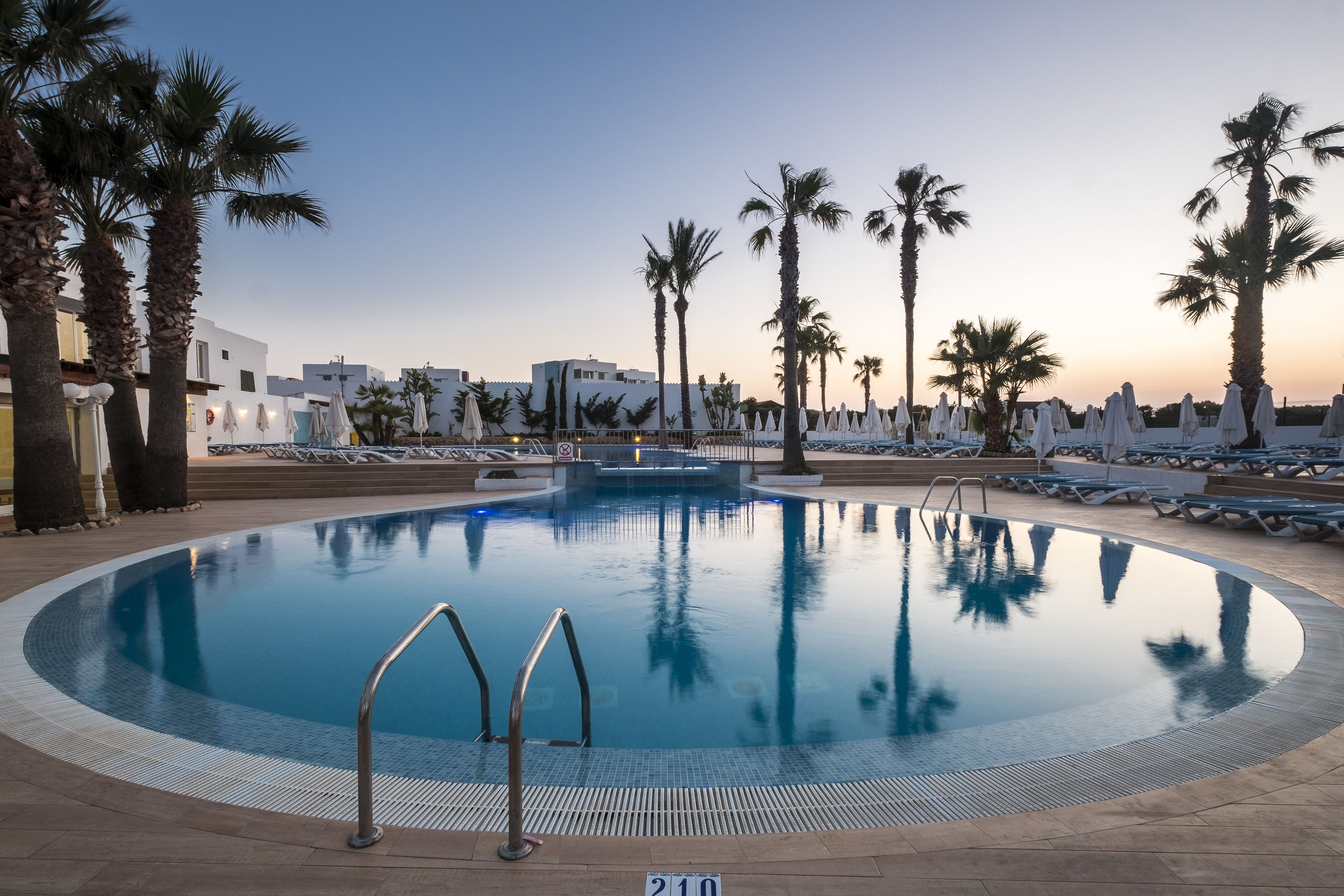 Una piscina con sillas de descanso y sombrillas, rodeada de palmeras y edificios.
