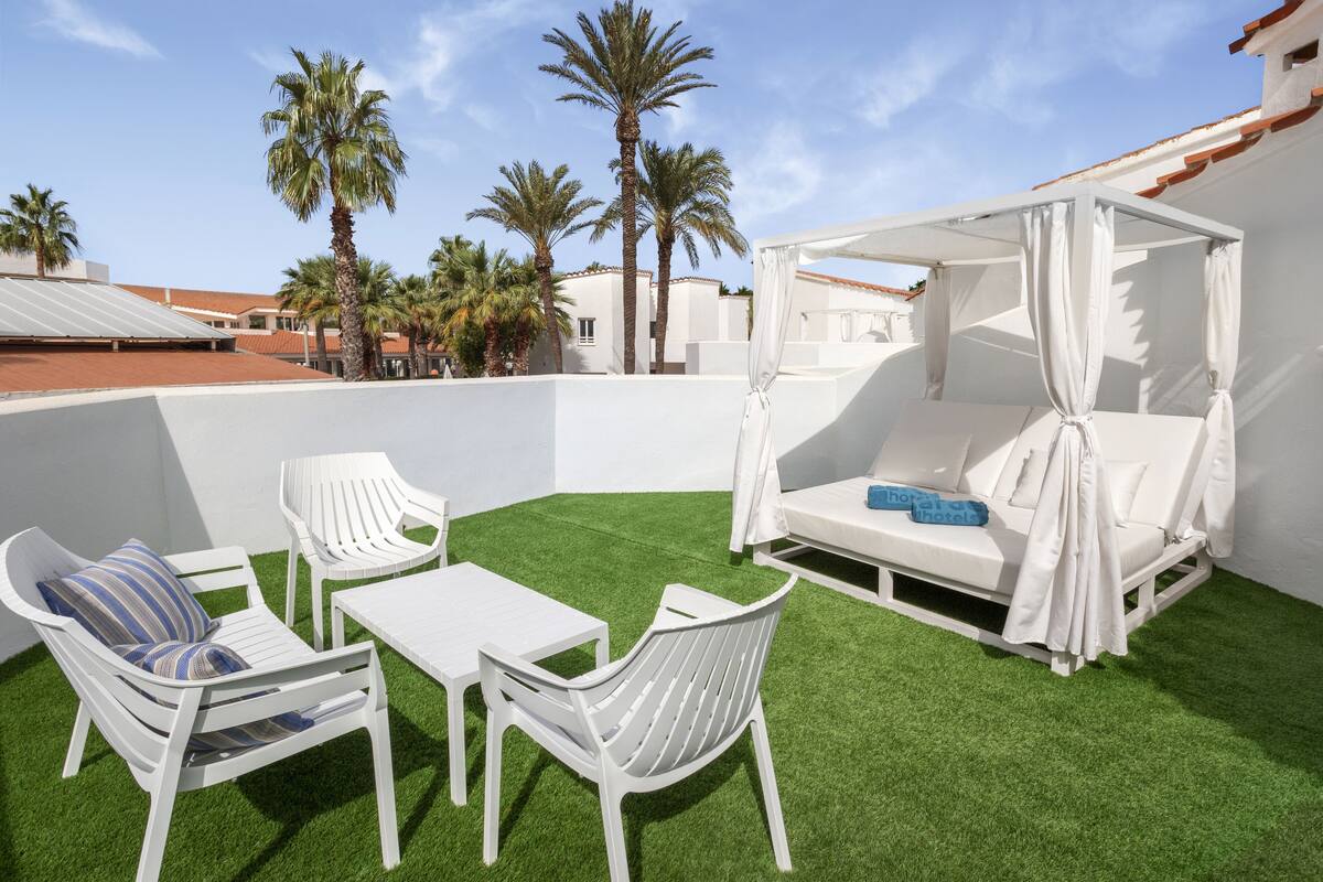 A rooftop terrace with white furniture, a canopy bed, and palm trees in the background.