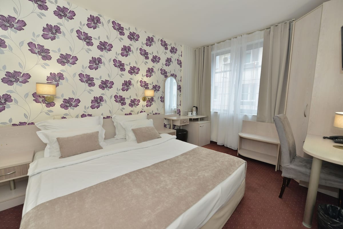 A hotel room with a large bed, a desk, and a chair. There is a mirror above the desk and a window with curtains.