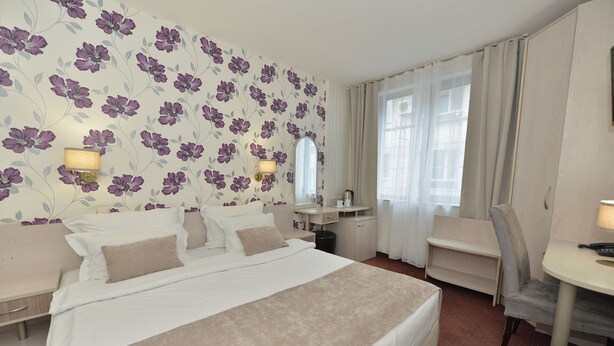 A hotel room with a large bed, a desk, and a chair. There is a mirror above the desk and a window with curtains.