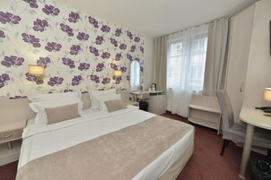 A hotel room with a large bed, a desk, and a chair. There is a mirror above the desk and a window with curtains.