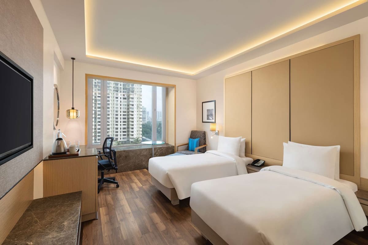 A hotel room with two beds, a desk with a chair, a TV, a window with a city view, and a bedside table with a lamp.