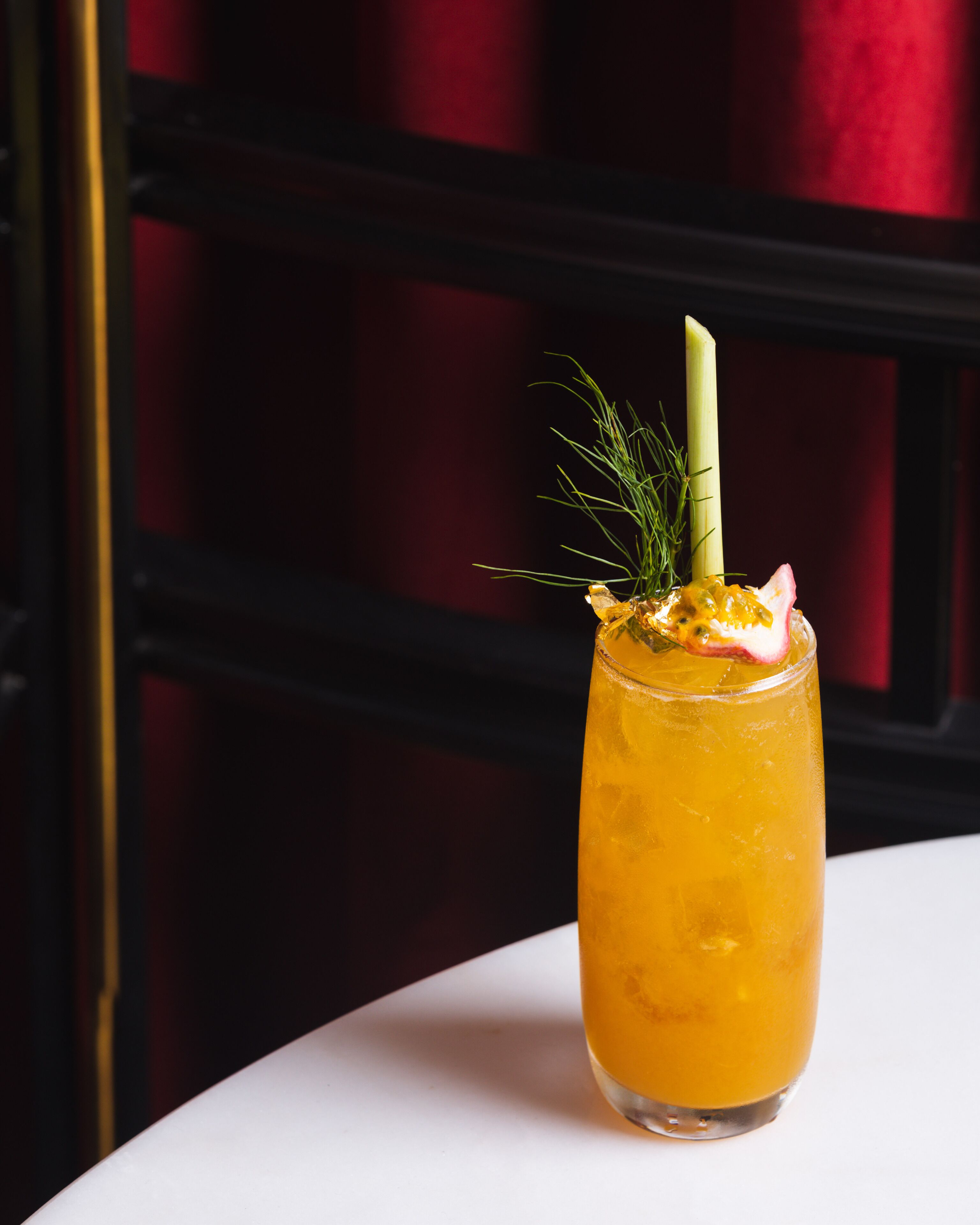 A tall glass with a yellow beverage, garnished with a sprig of greenery and a slice of fruit, placed on a white surface against a dark background.