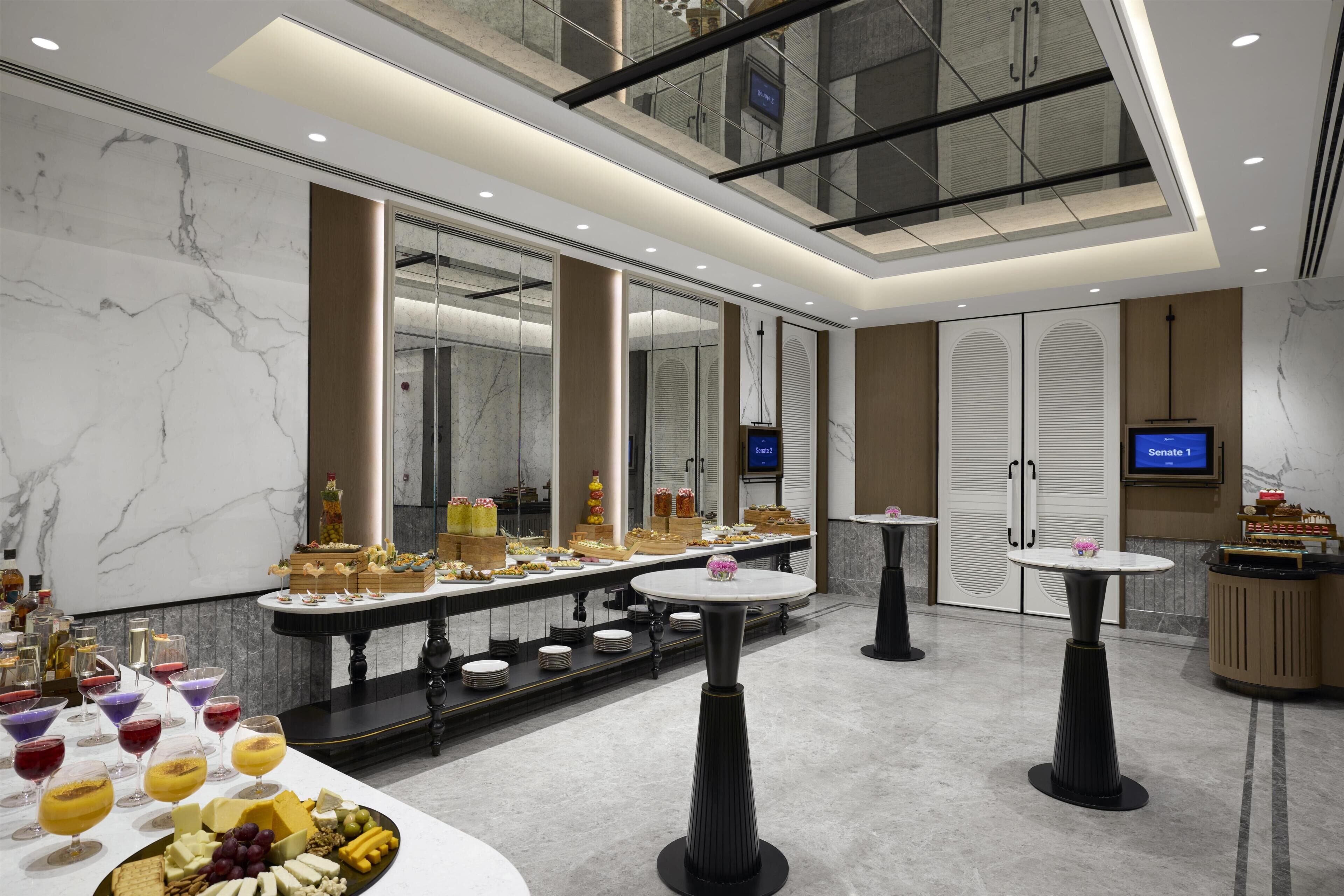 Un hall d’hôtel moderne avec des murs en marbre, un plafond en verre et un espace buffet proposant une variété de plats et de boissons.