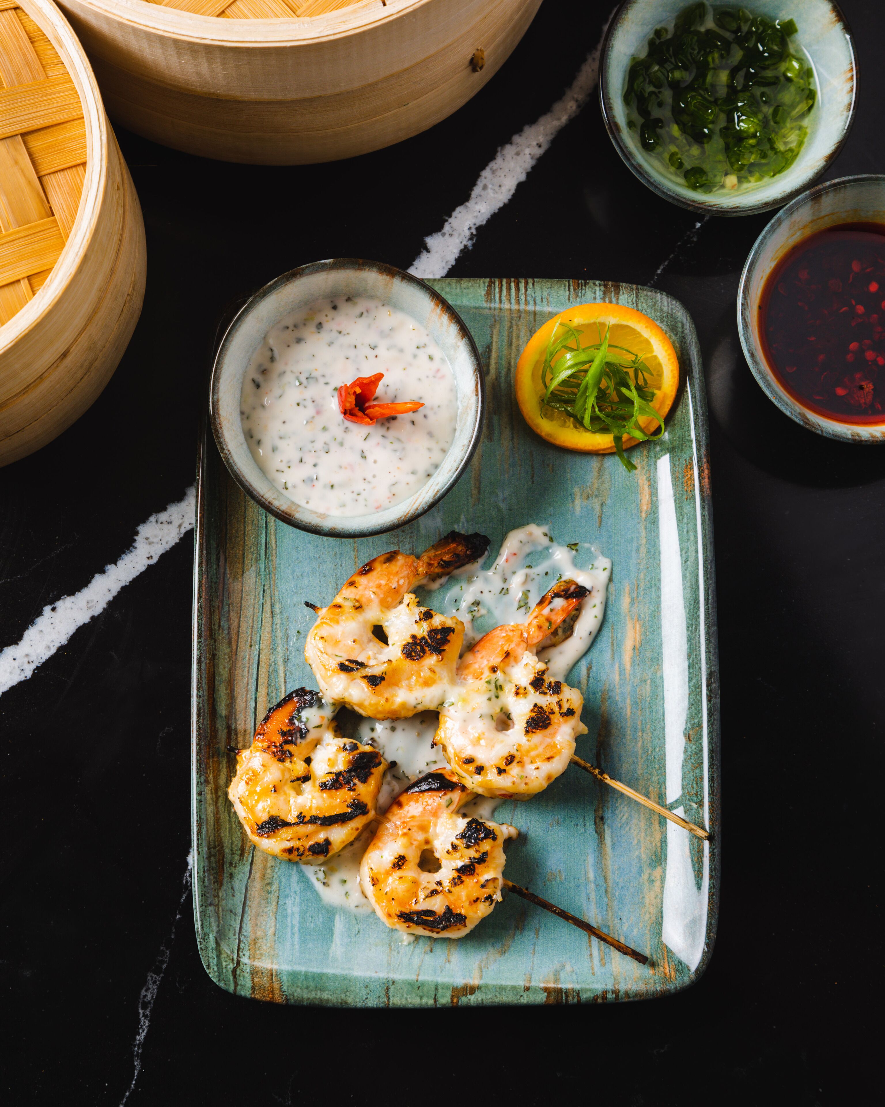 A plate with grilled chicken skewers, a bowl of sauce, and a small bowl of greens.