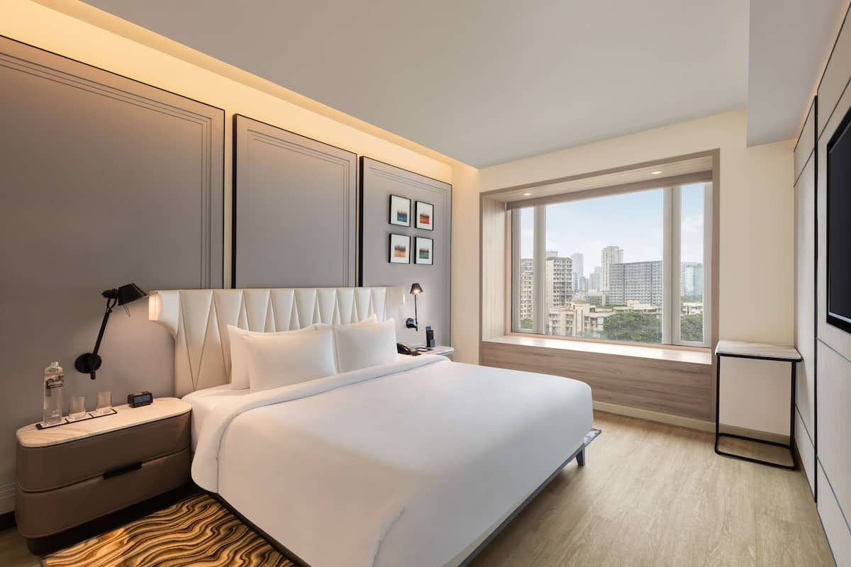 A modern hotel room with a large bed, a nightstand, a TV, and a city view.
