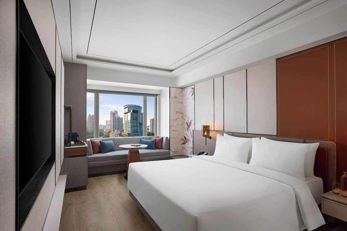 A hotel room with a large bed, a sofa, a TV, a city view, and a window.
