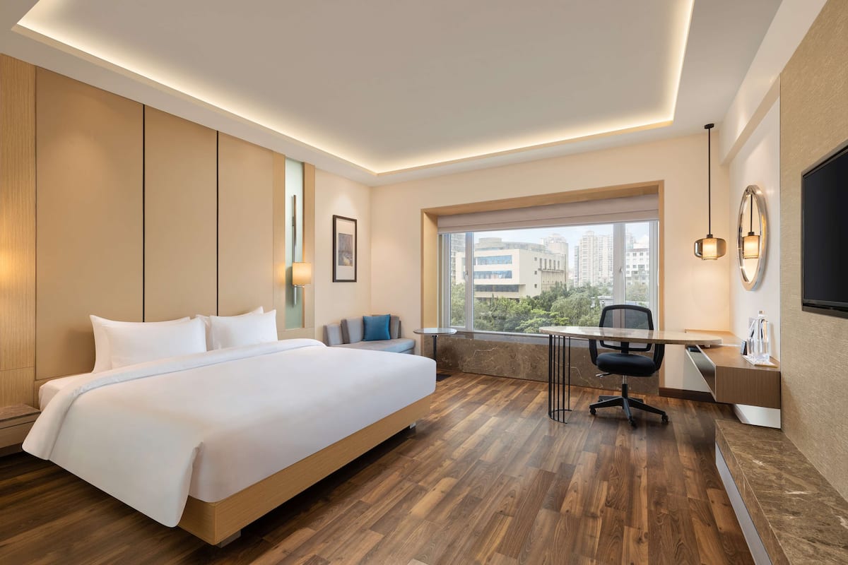 A modern hotel room with a large bed, a desk, and a view of the city through a large window.