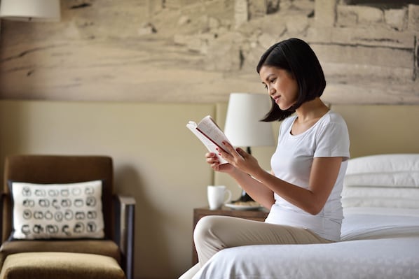 Una persona sentada en una cama leyendo un libro en una habitación con pared de piedra, una lámpara y un cojín estampado.