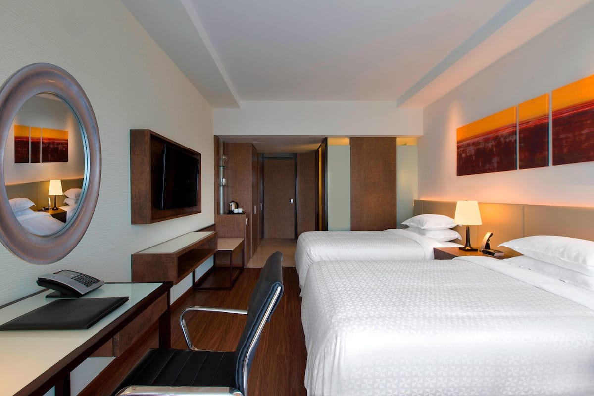 A hotel room with a desk, chair, two beds, a TV, and artwork on the wall.