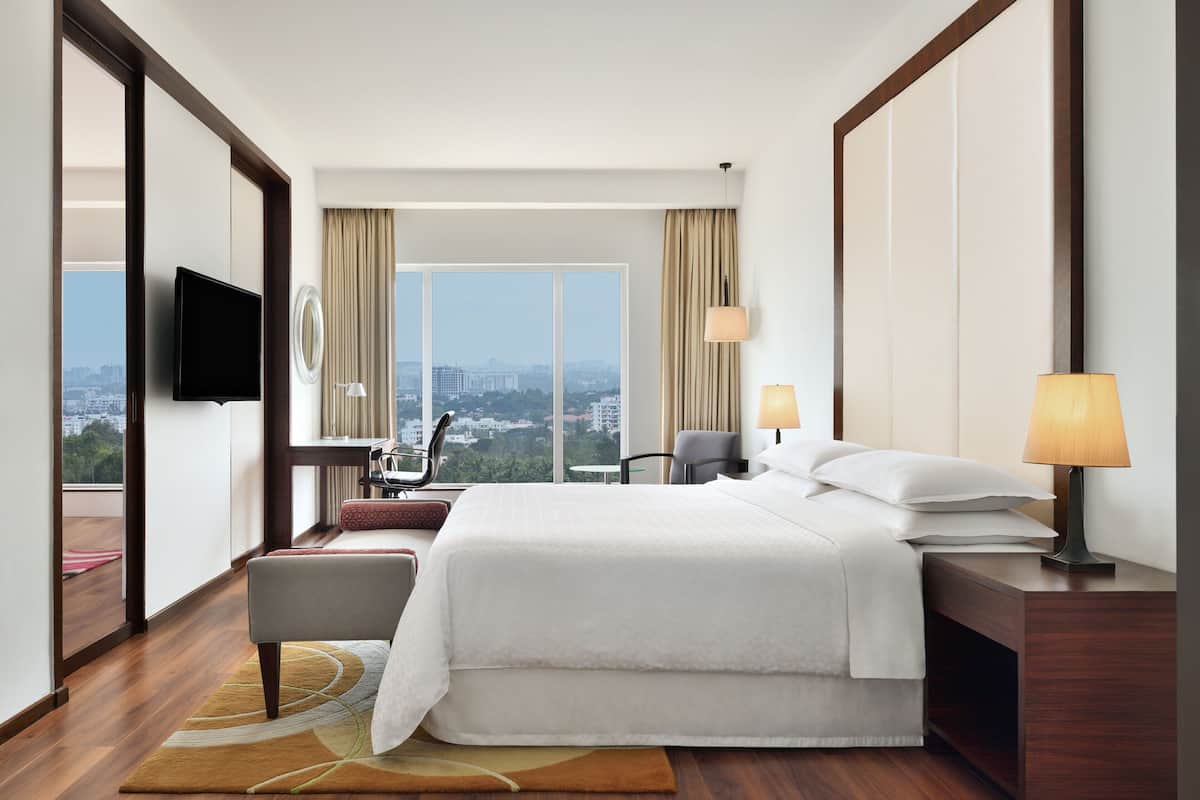 A hotel room with a large bed, a TV, a desk with a chair, and a view of the city.