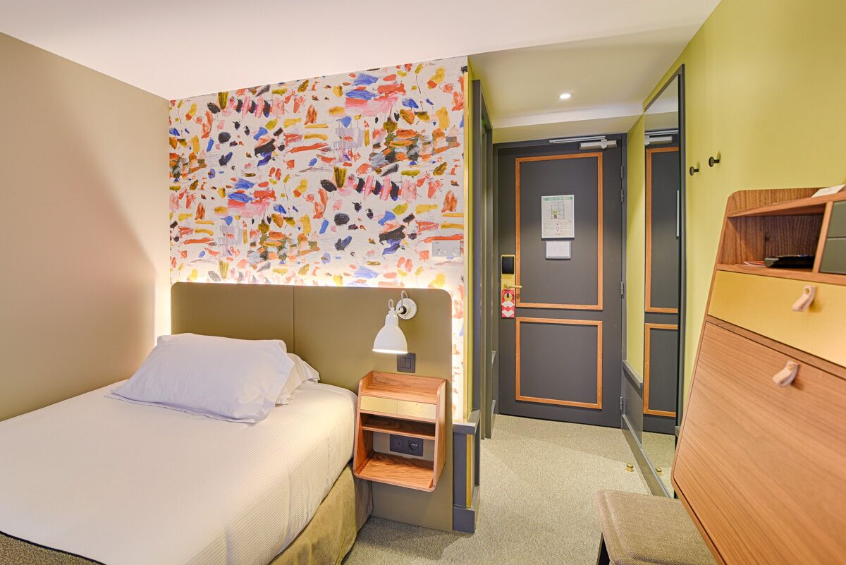 A hotel room with a bed, bedside table, lamp, and a colorful abstract wall.