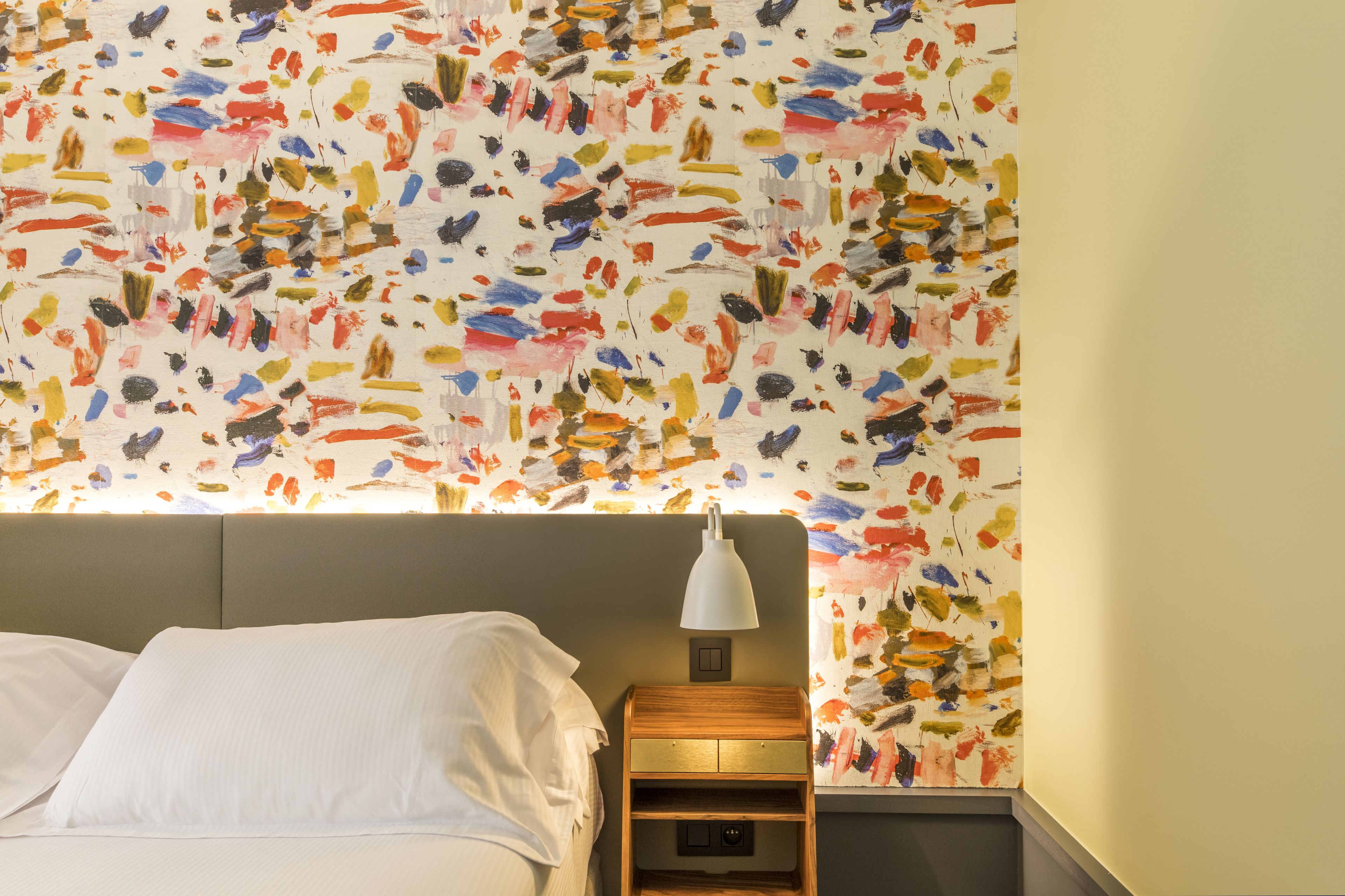 A hotel room with a bed, a nightstand, a lamp, and a wall with a colorful abstract pattern.