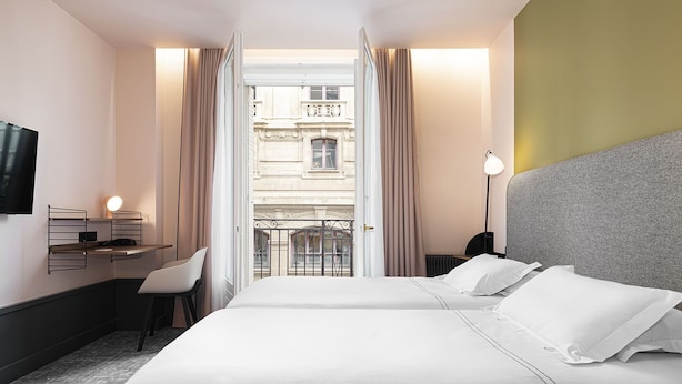 Una habitación de hotel con una cama grande, un escritorio con silla, un televisor en la pared y vistas a un edificio a través de una ventana abierta.