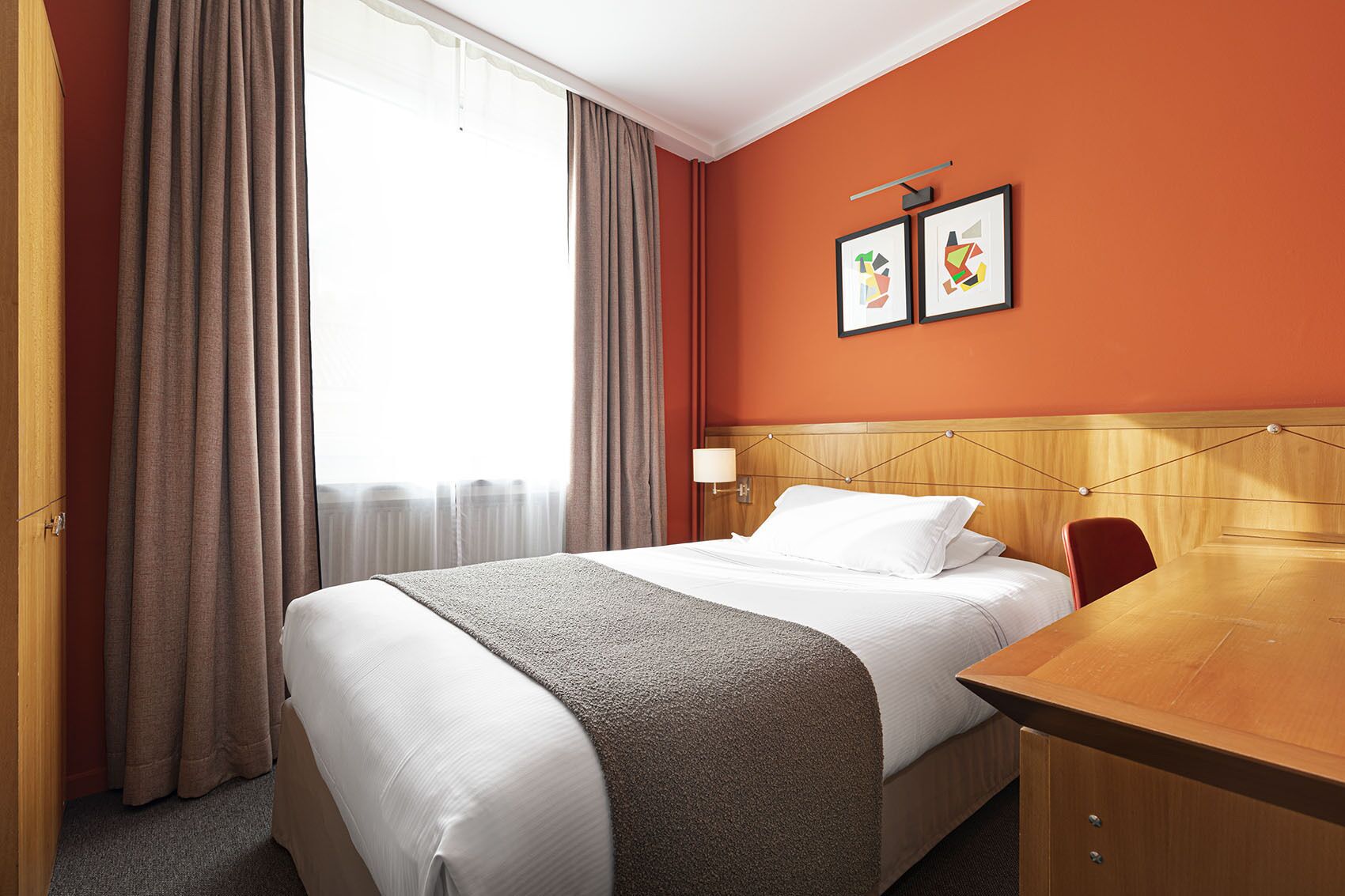 Una camera d'albergo con un letto, una testiera in legno, una scrivania e una sedia. Alle finestre ci sono tende e due quadri appesi al muro.