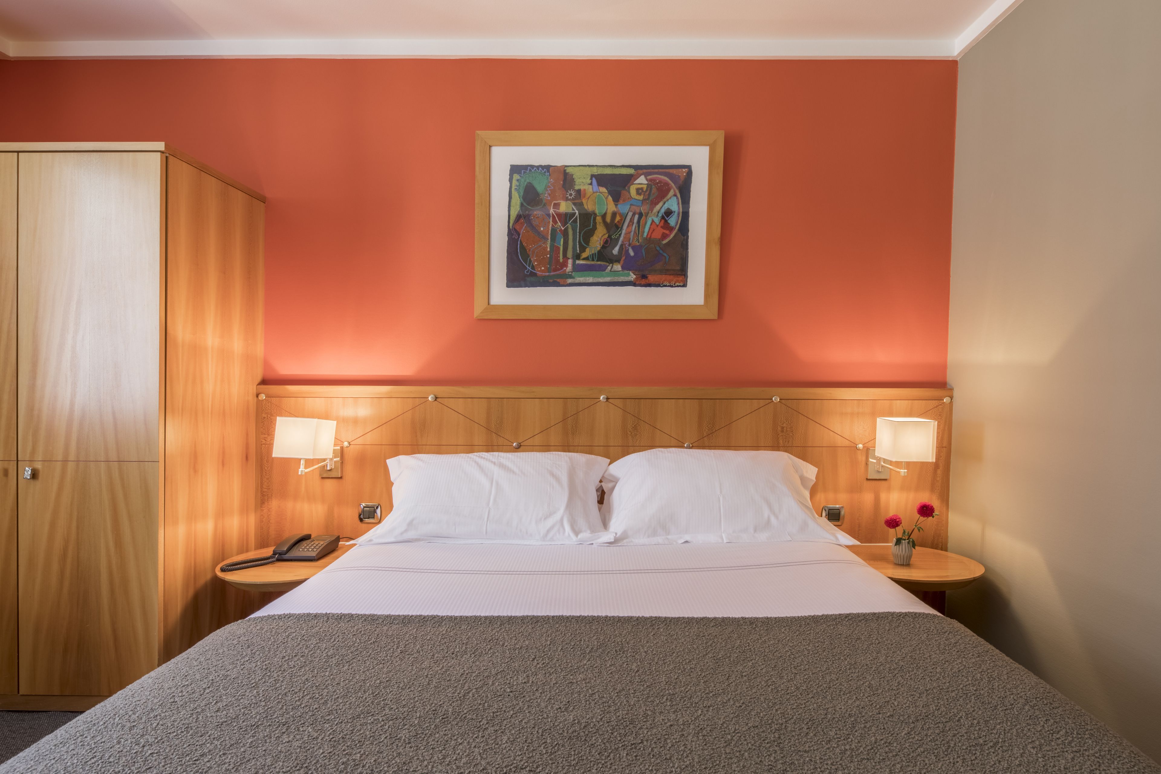 A hotel room with a bed, bedside lamps, a wooden wardrobe, a painting on the wall, and a small table with flowers.