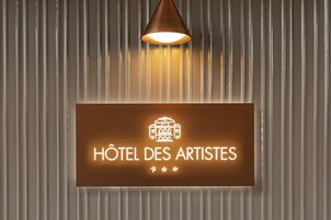 Un cartello con su scritto "Hôtel des Artistes" e un'icona di hotel, fissato a una parete in lamiera ondulata.